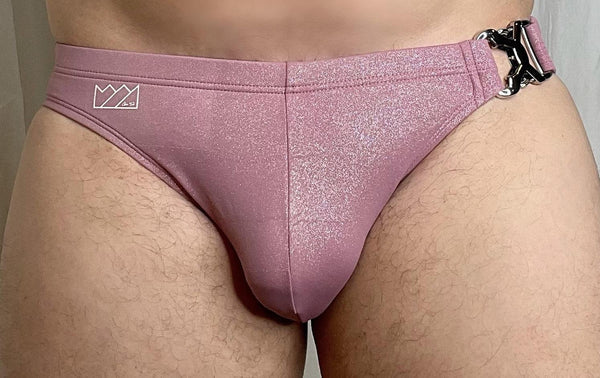 A shimmering rose gold glittery swim brief with a black metal clasp and white logo patch for summer wear