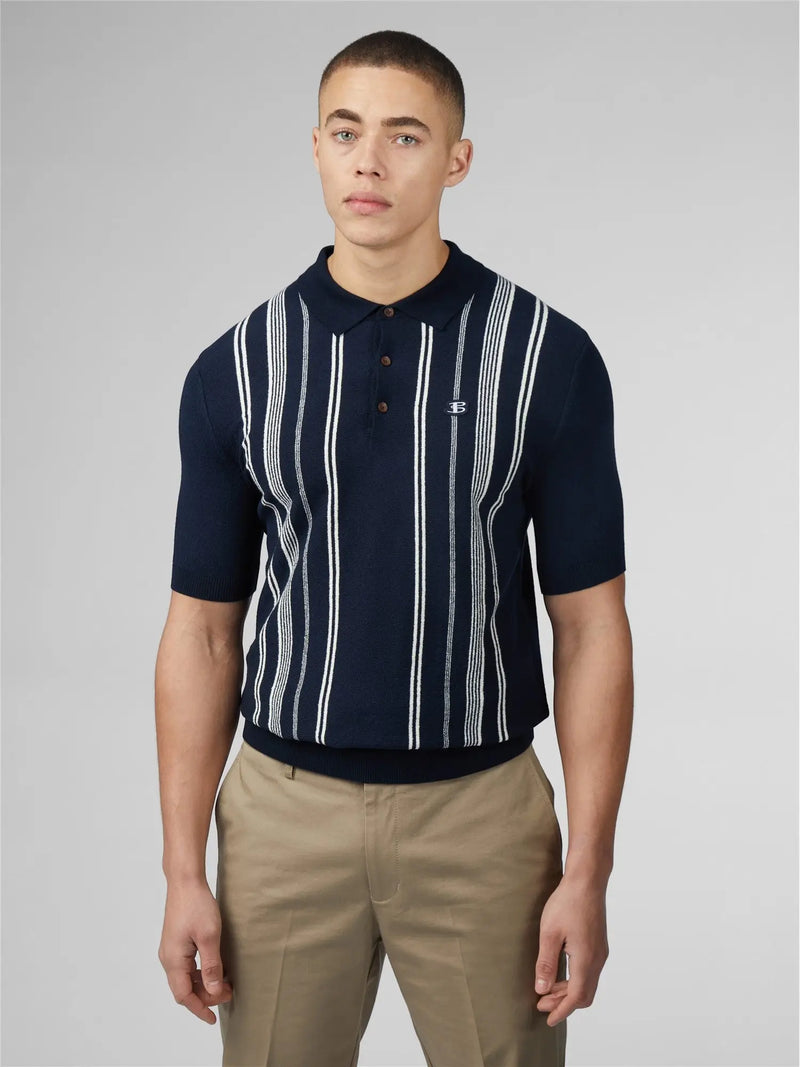 A man wears a navy blue vertically striped polo shirt with khaki pants.