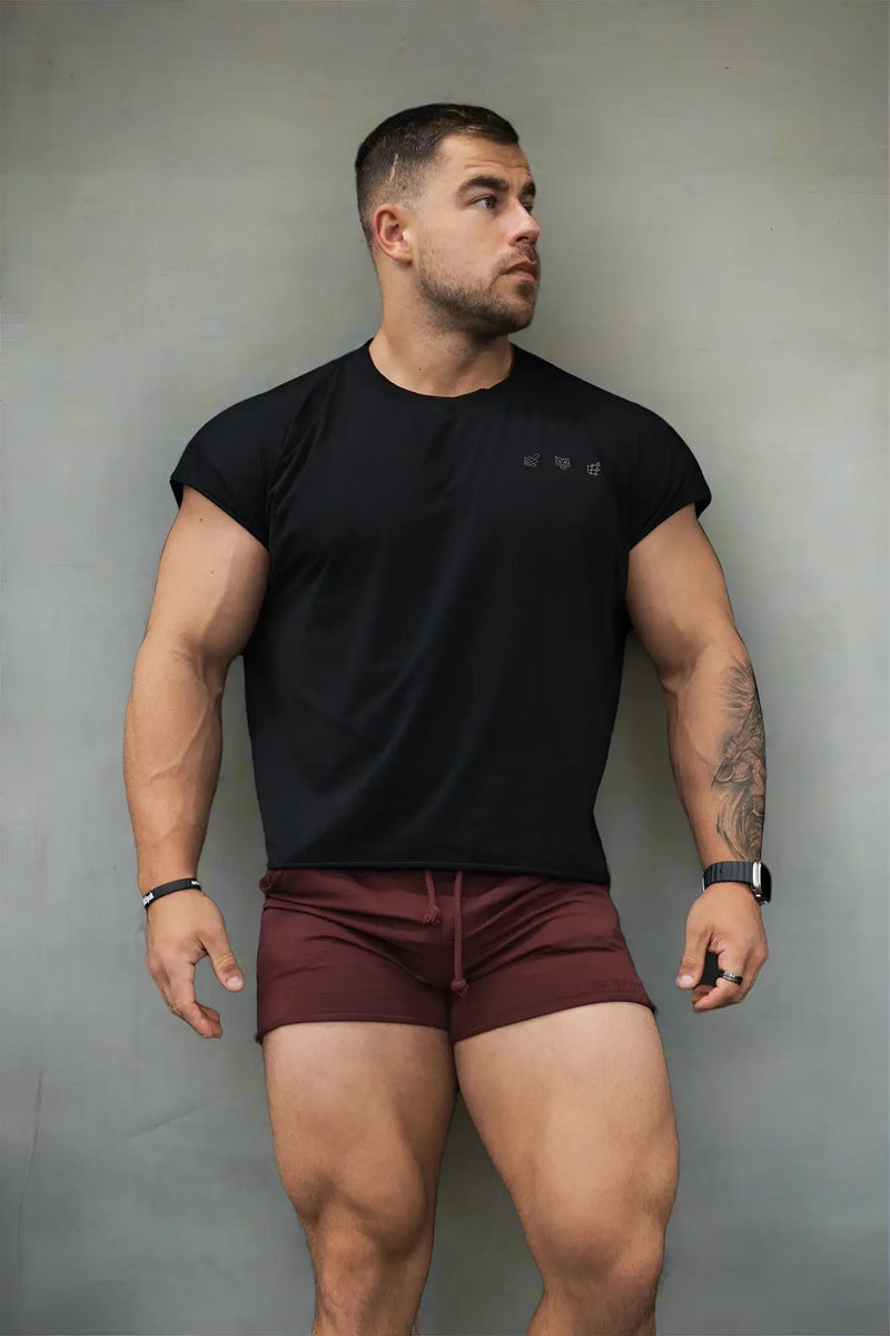 Muscular man wears black crewneck shirt and maroon shorts against gray wall.