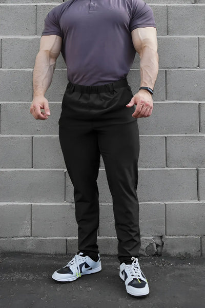 Muscular man wearing gray polo and black athletic pants against gray bricks.