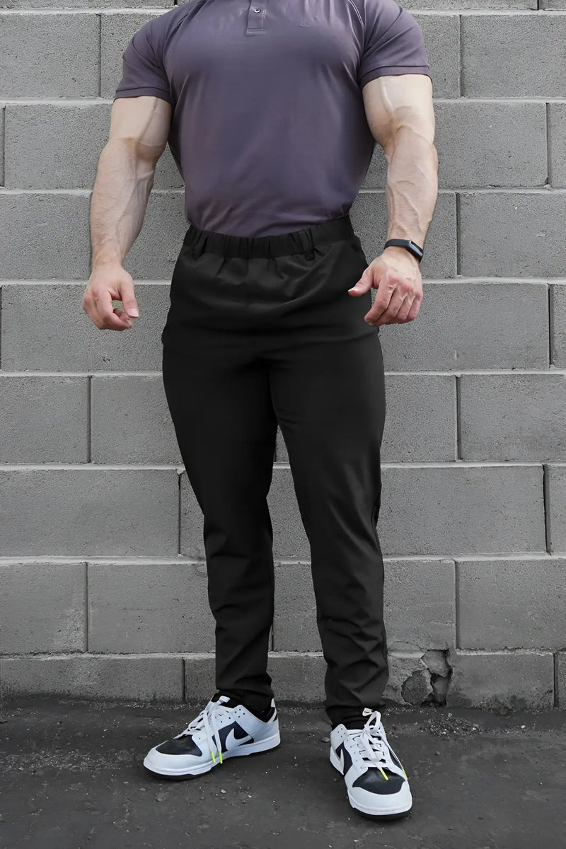 Muscular man wearing gray polo and black athletic pants against gray bricks.
