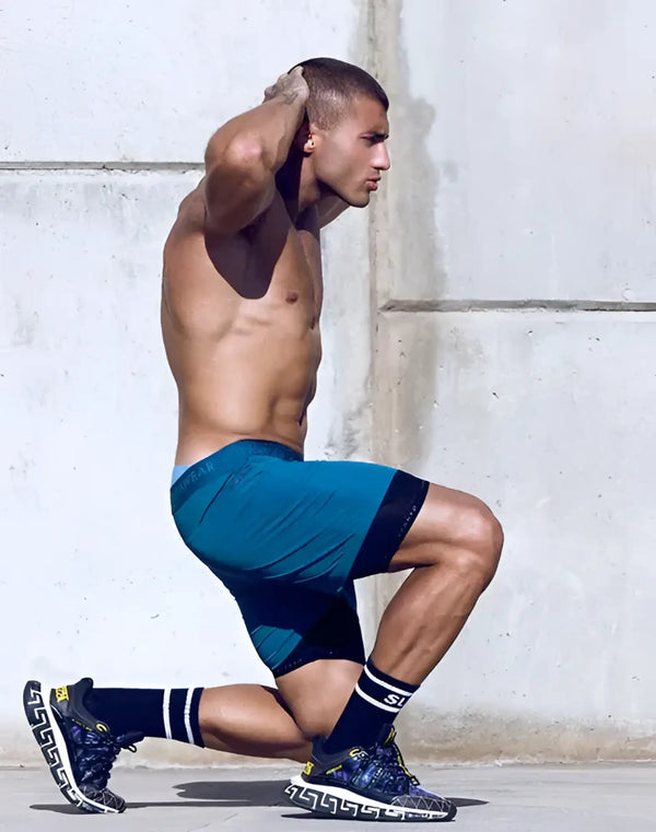 Shirtless man in blue athletic shorts and black sneakers kneels outdoors.