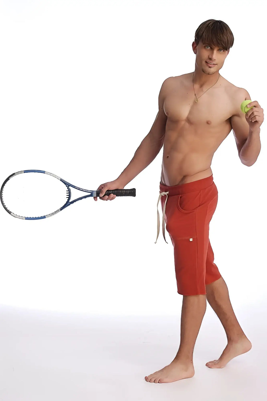 A man wearing cinnabar eco-track shorts with white drawstrings and gold logo holding a blue racket and yellow ball