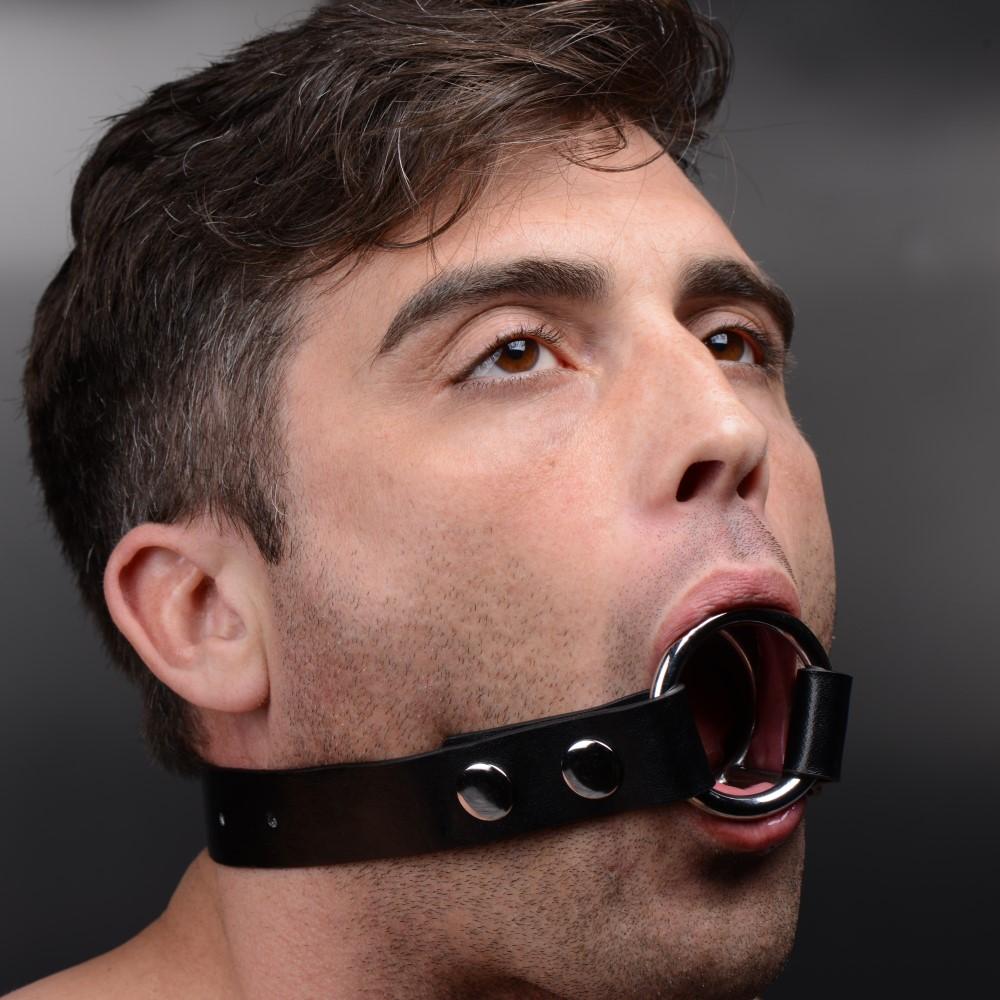 Strict The Deep Throat Gag: A man wearing a black leather neck strap featuring a silver metal O-ring gag in his mouth.