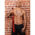 Strict Leather The Heavy Tail Flogger with red and black leather straps held by a shirtless man against a brick wall.