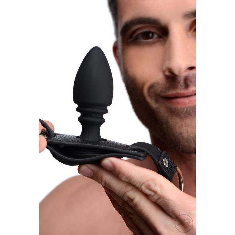 A man holds a black Strict silicone anal plug attached to a matching leather-textured cock ring harness.