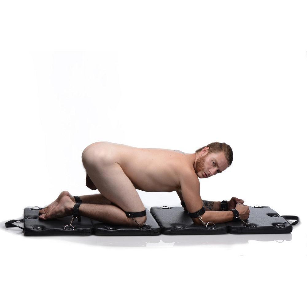 Strict XL Bondage Board: A man on hands and knees, limbs secured to a foldable, black padded mat with metal rings.