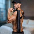 A man wearing a leather harness holds a black Strict Leather flogger with a polished stainless steel handle.