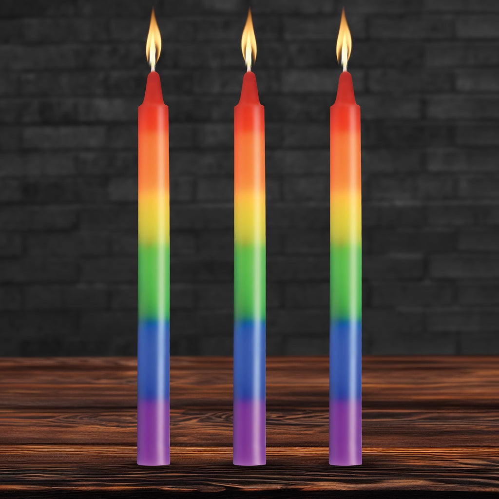 Three Master Series Pride taper candles in rainbow stripes, each with a lit flame, standing on a wooden surface.