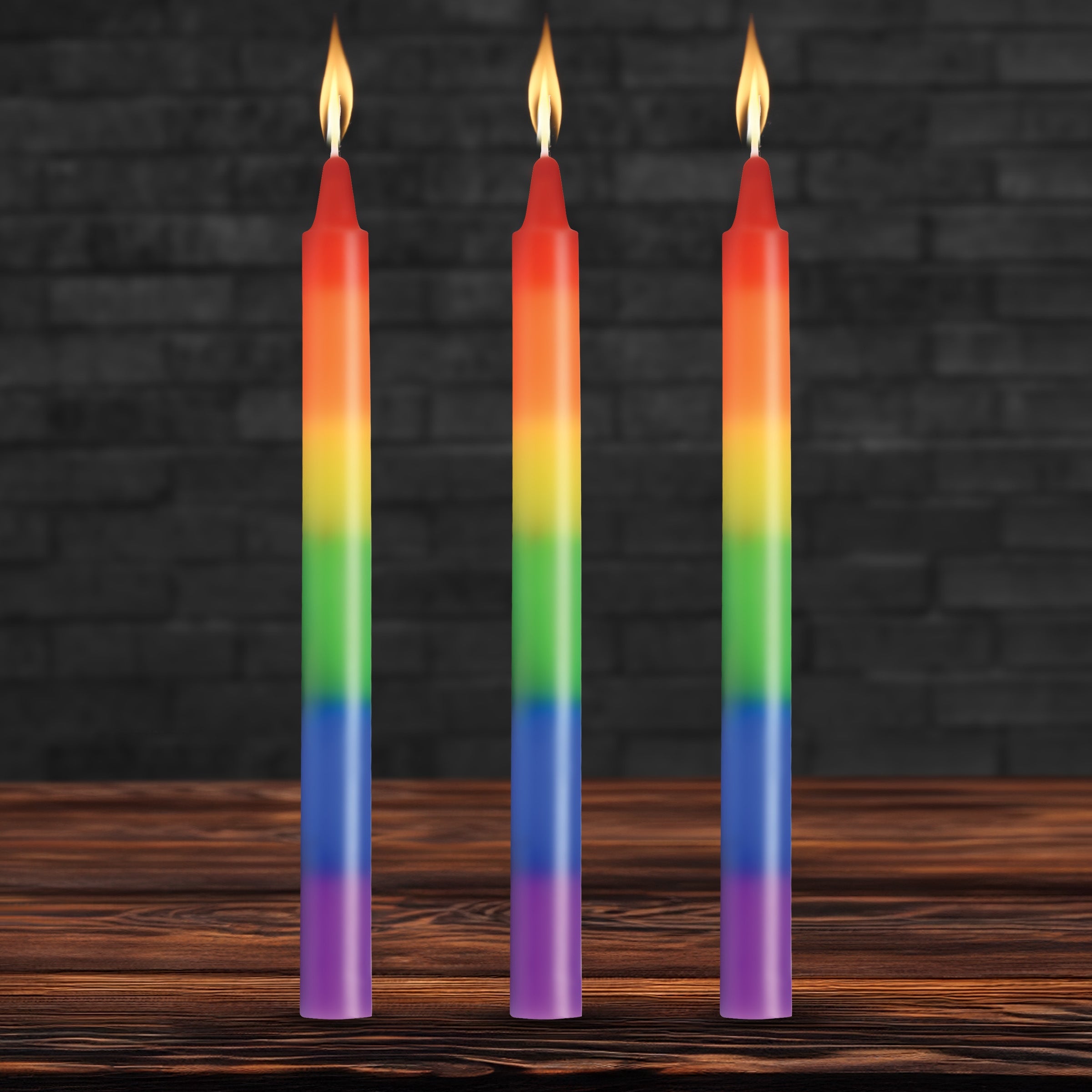 Three Master Series Pride taper candles in rainbow stripes, each with a lit flame, standing on a wooden surface.