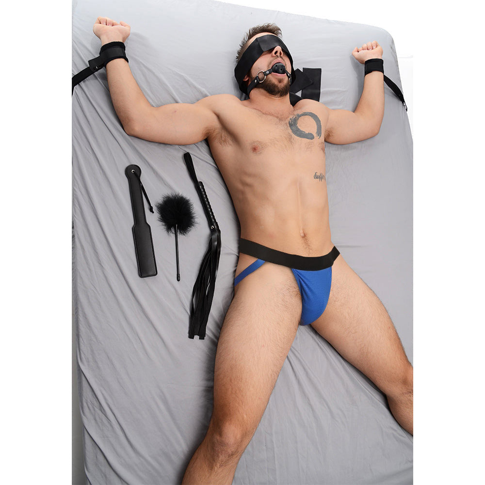 Strict Bed Restraint Bondage Kit featuring a man in black restraints, blindfold, ball gag, and assorted black accessories.