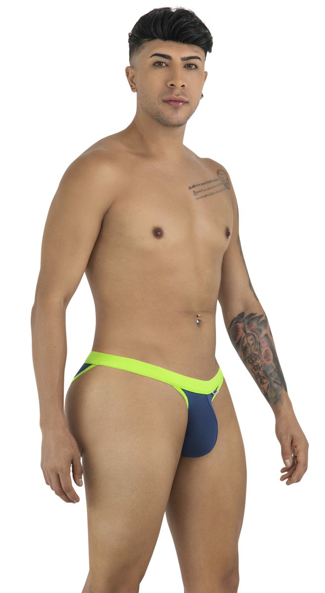 Man standing in profile wearing a navy and lime green bikini, showing arm and chest tattoos against a white background.