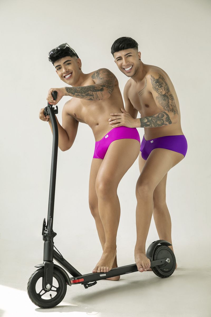 Two shirtless, tattooed men wearing orchid trunks pose together on a black electric scooter against a white background.