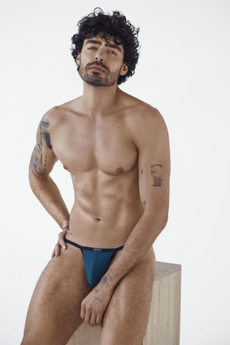 A model sits on a block wearing petrol blue Boreas briefs with thin straps.
