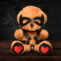 A plush tan dom teddy bear wearing a black leather mask and harness with red heart patches on its paws