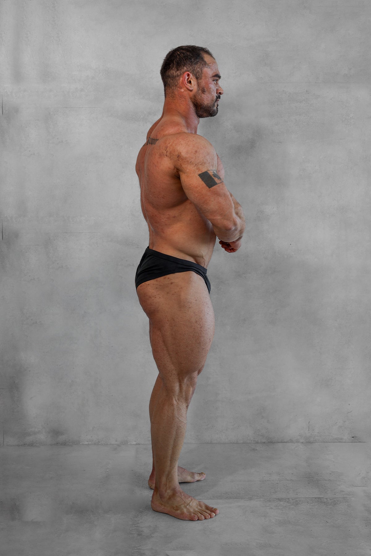 A muscular man stands in profile wearing black classic physique trunks 2 inch seam designed for men’s fitness