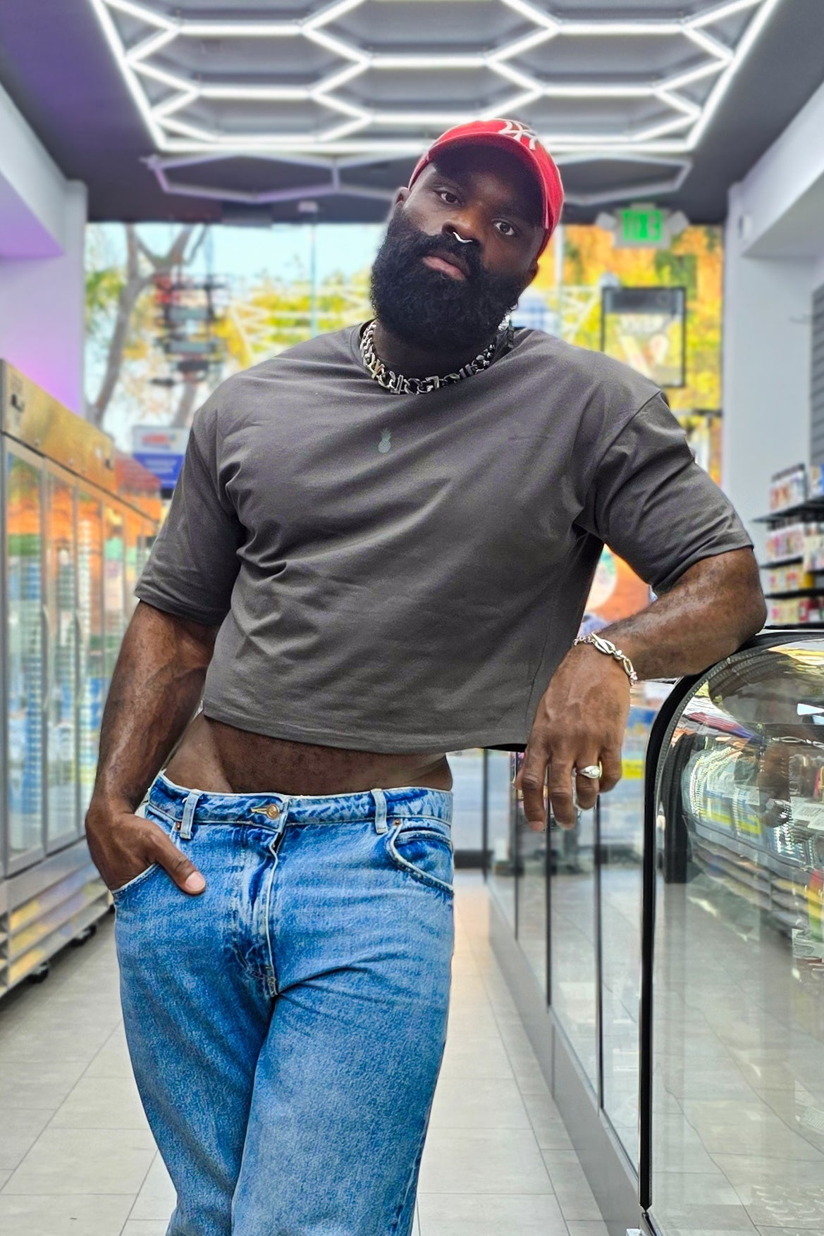 A man wearing a dark grey oversized relaxed crop top with denim jeans and a red baseball cap facing forward