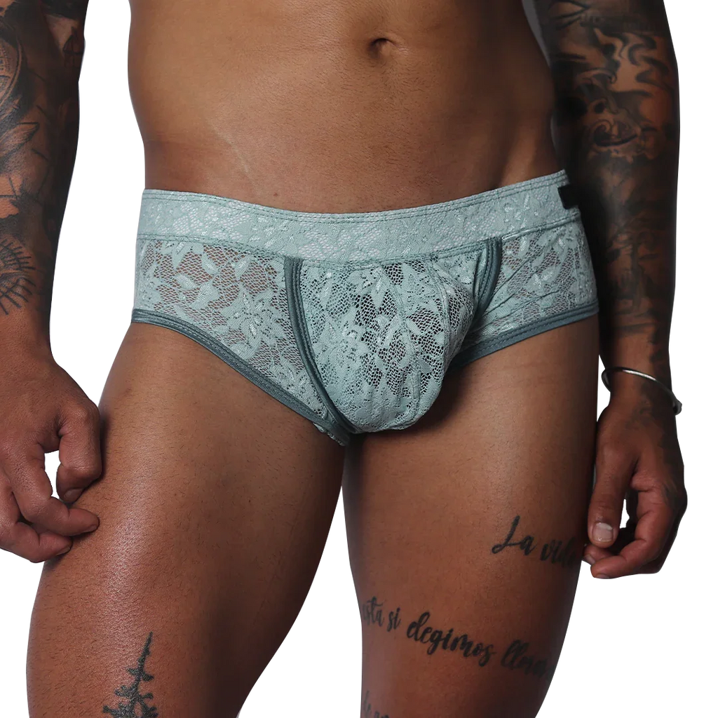 Aqua blue floral lace trunk with solid piping and a contoured pouch, modeled on a tattooed torso.