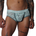 Aqua blue floral lace trunk with solid piping and a contoured pouch, modeled on a tattooed torso.