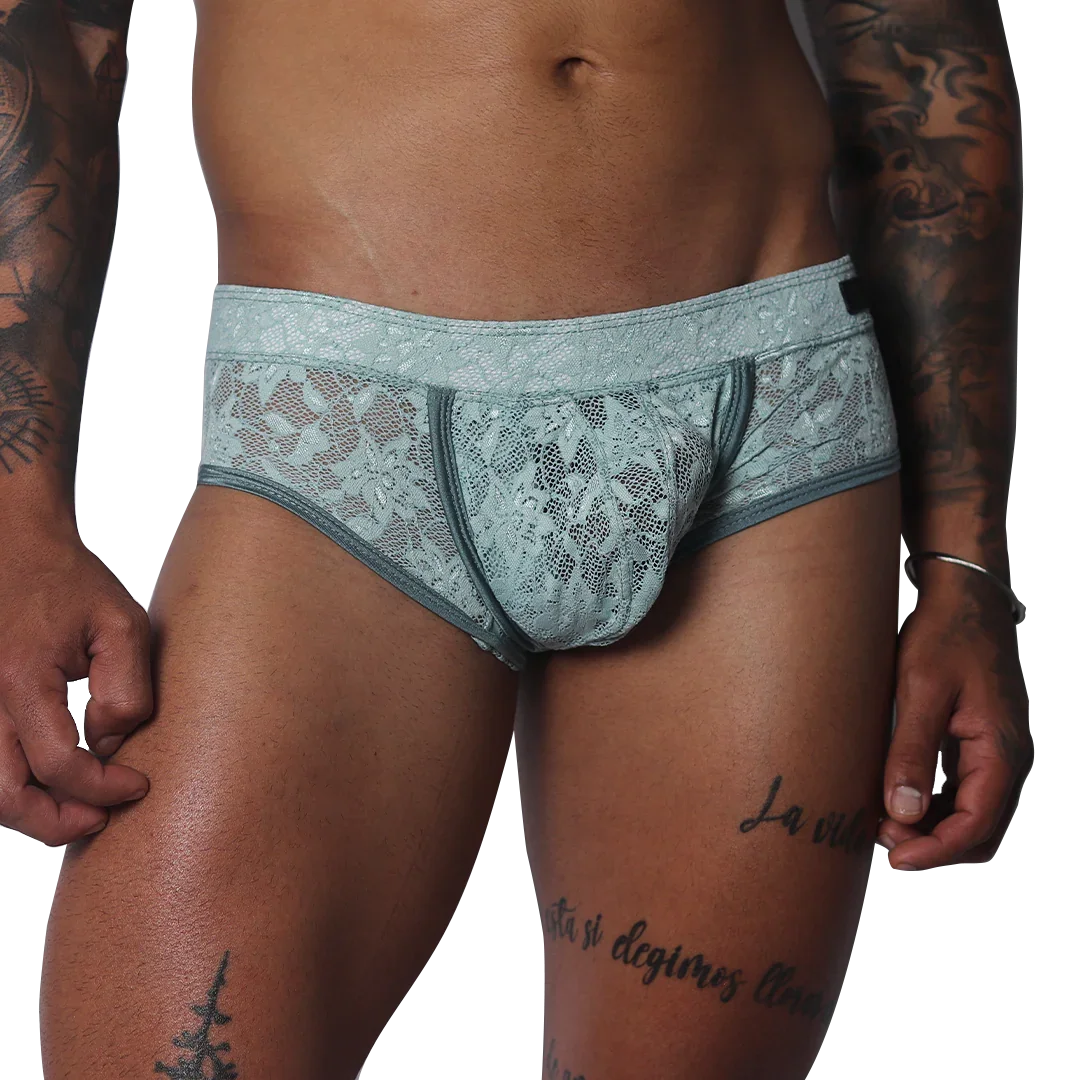 Aqua blue floral lace trunk with solid piping and a contoured pouch, modeled on a tattooed torso.