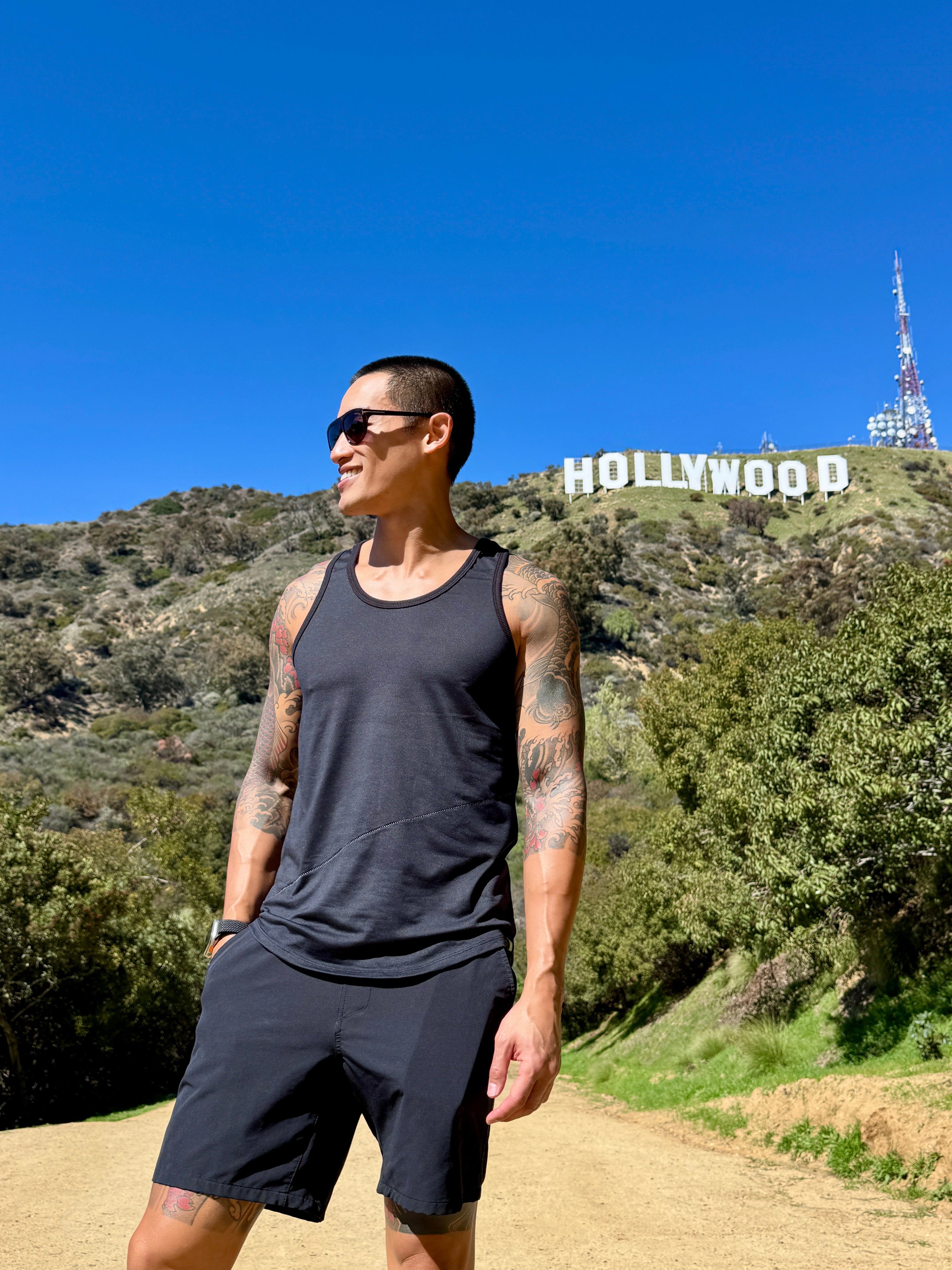 A man stands on a dirt path wearing the perfect tank black carbon sleeveless top and matching shorts with hollywood sign background