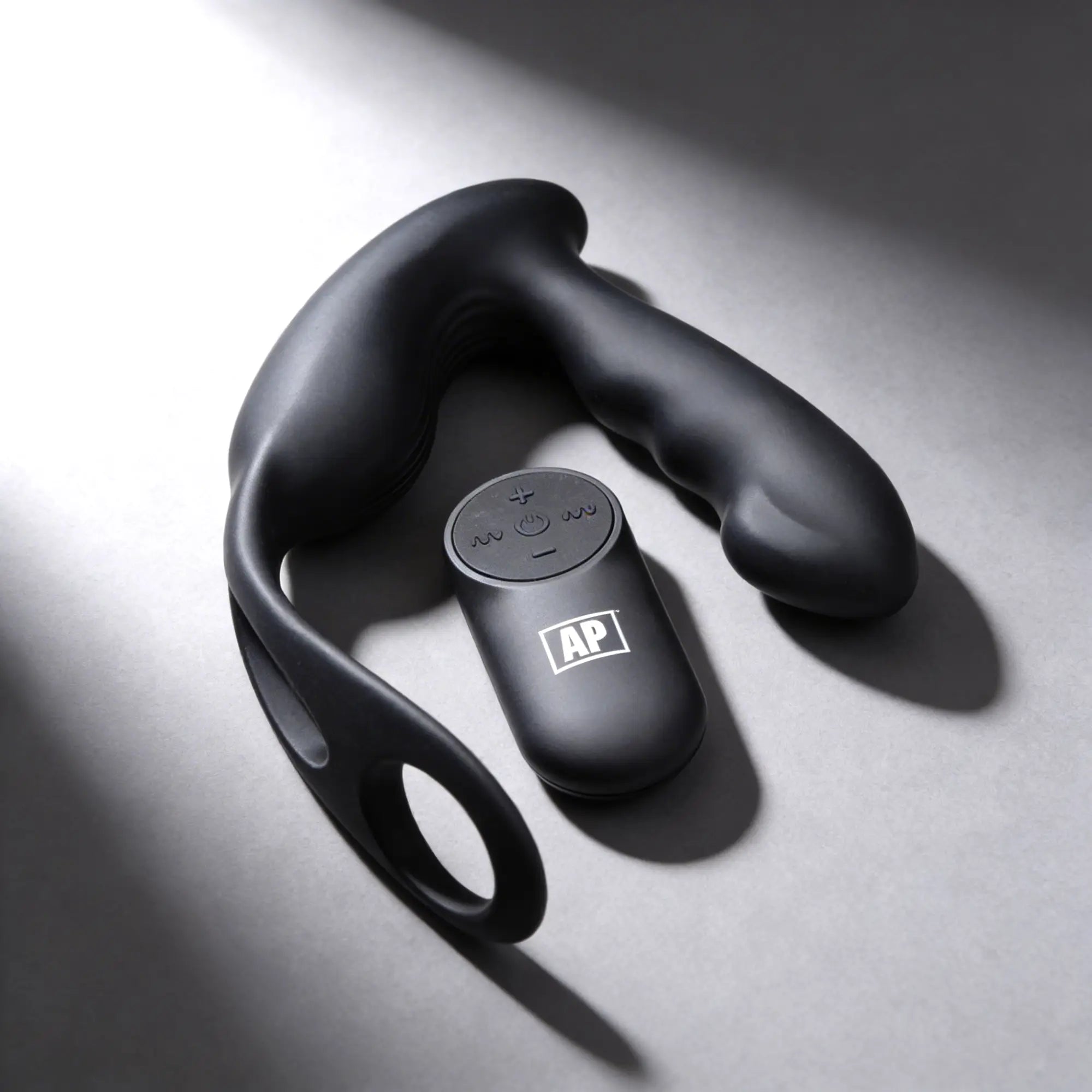 Alpha Pro 7X black silicone prostate stimulator and harness with a matching remote control on a grey surface.
