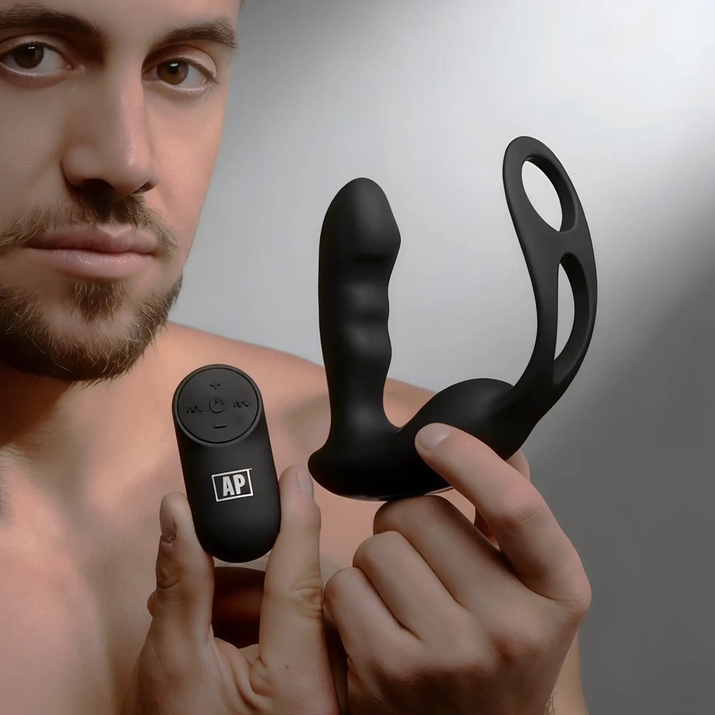 Alpha Pro 7X black silicone prostate stimulator with a cock and ball harness and remote control held by a man.