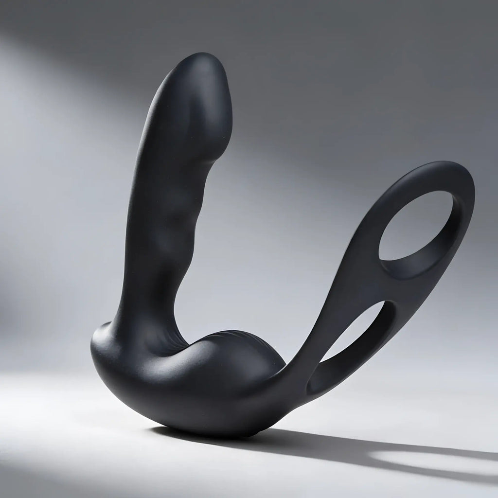 Matte black Alpha Pro 7X prostate stimulator with curved shaft and integrated cock and ball harness against a grey background.