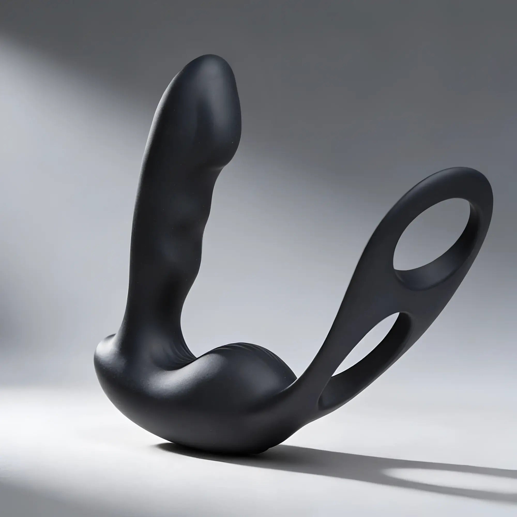 Matte black Alpha Pro 7X prostate stimulator with curved shaft and integrated cock and ball harness against a grey background.