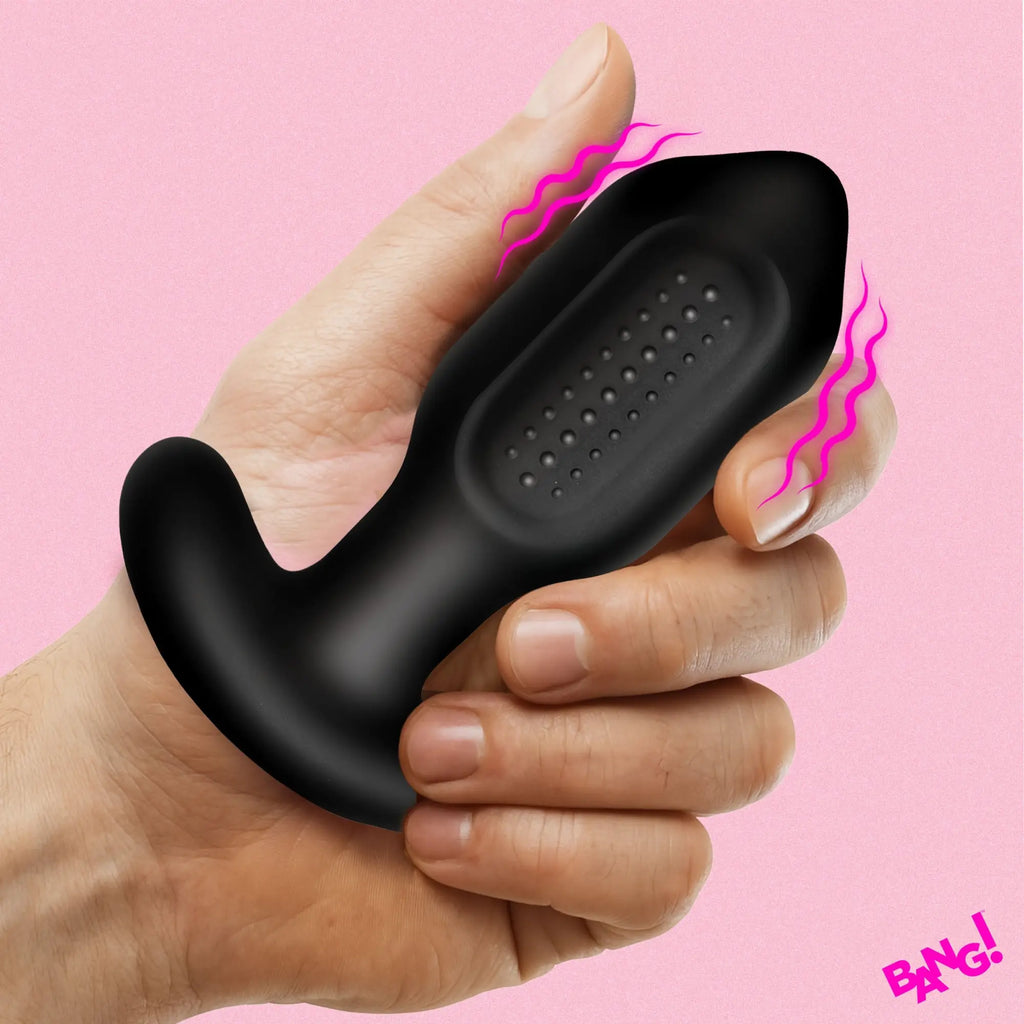 A hand holds a black, silicone Bang Bumpy Vibrating Butt Plug with a textured, dotted surface against a pink background.