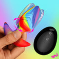 Hand holding a rainbow-colored Bang rotating, vibrating silicone butt plug next to a black remote control.