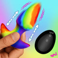 Bang Thrusting and Vibrating Silicone Butt Plug in a rainbow color pattern, held in hand with a black remote control.