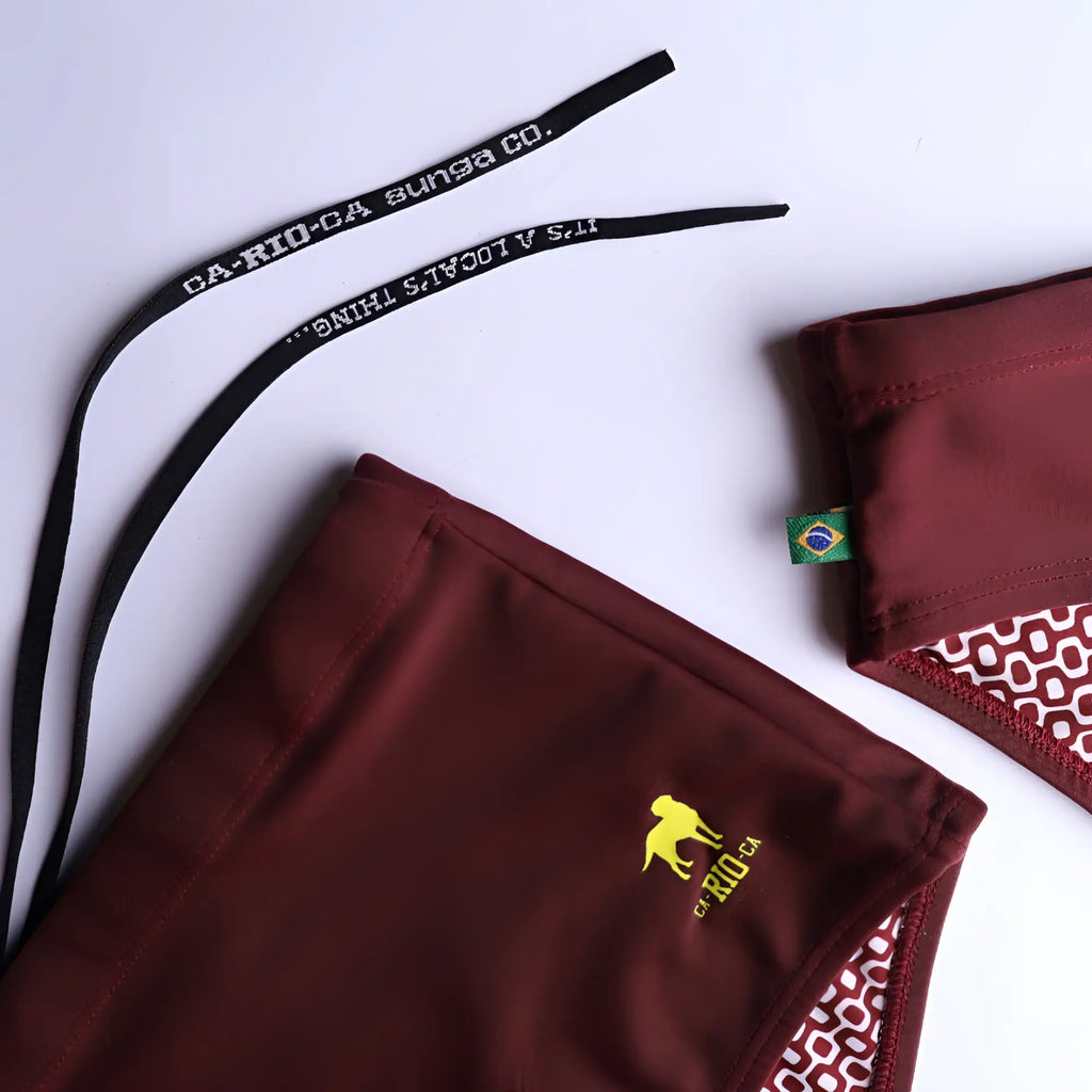 Burgundy CA-RIO-CA sunga swimming trunks with a yellow logo, patterned interior, and branded black drawstrings.