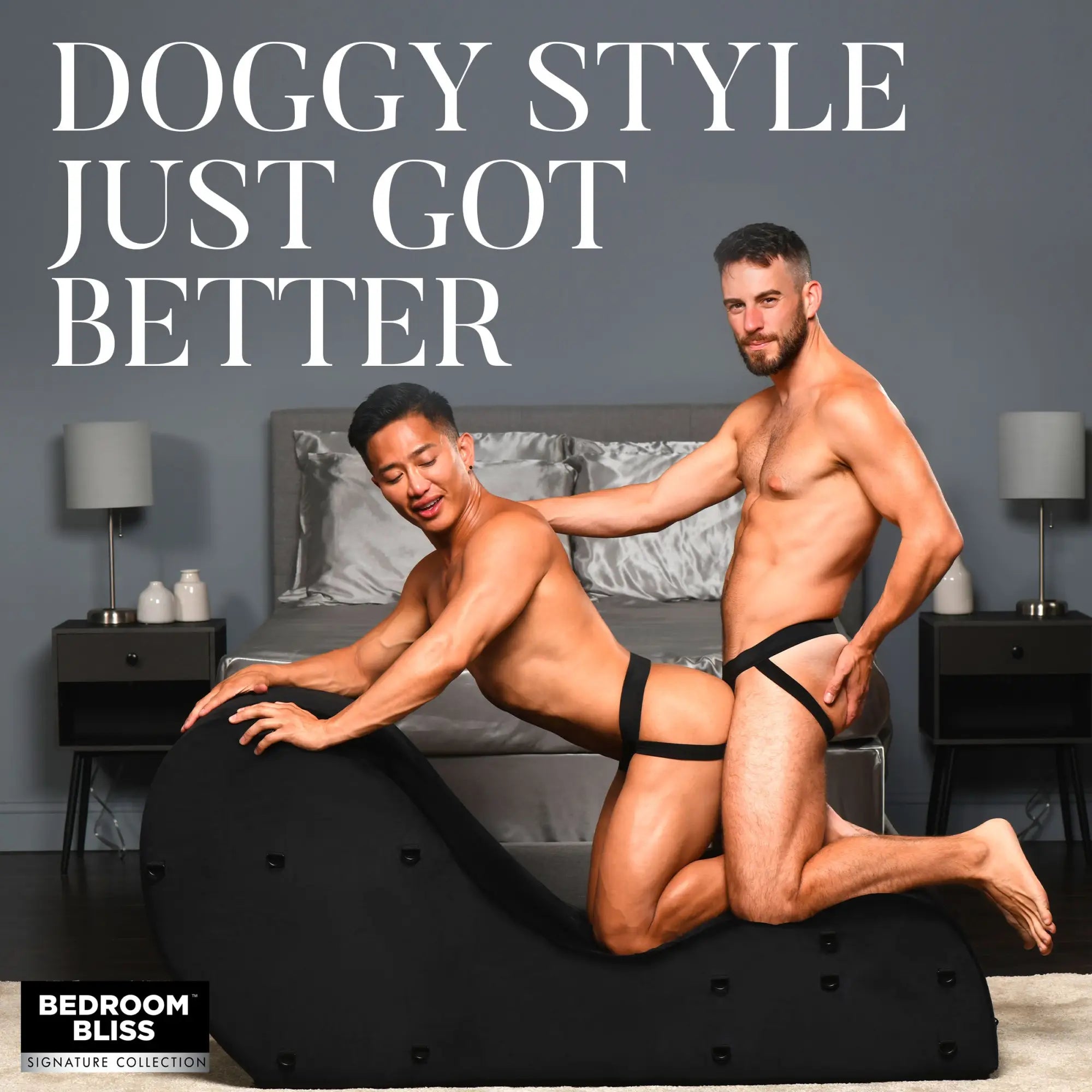 Two men in jockstraps pose with a black Bedroom Bliss Bondage Love Couch in a modern bedroom.