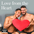 Two smiling men holding a red Bedroom Bliss Heart Pillow in a bedroom setting. Text reads, "Love from the Heart."