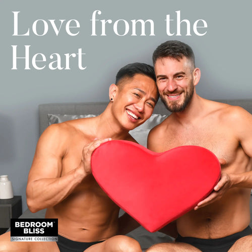 Two smiling men holding a red Bedroom Bliss Heart Pillow in a bedroom setting. Text reads, "Love from the Heart."