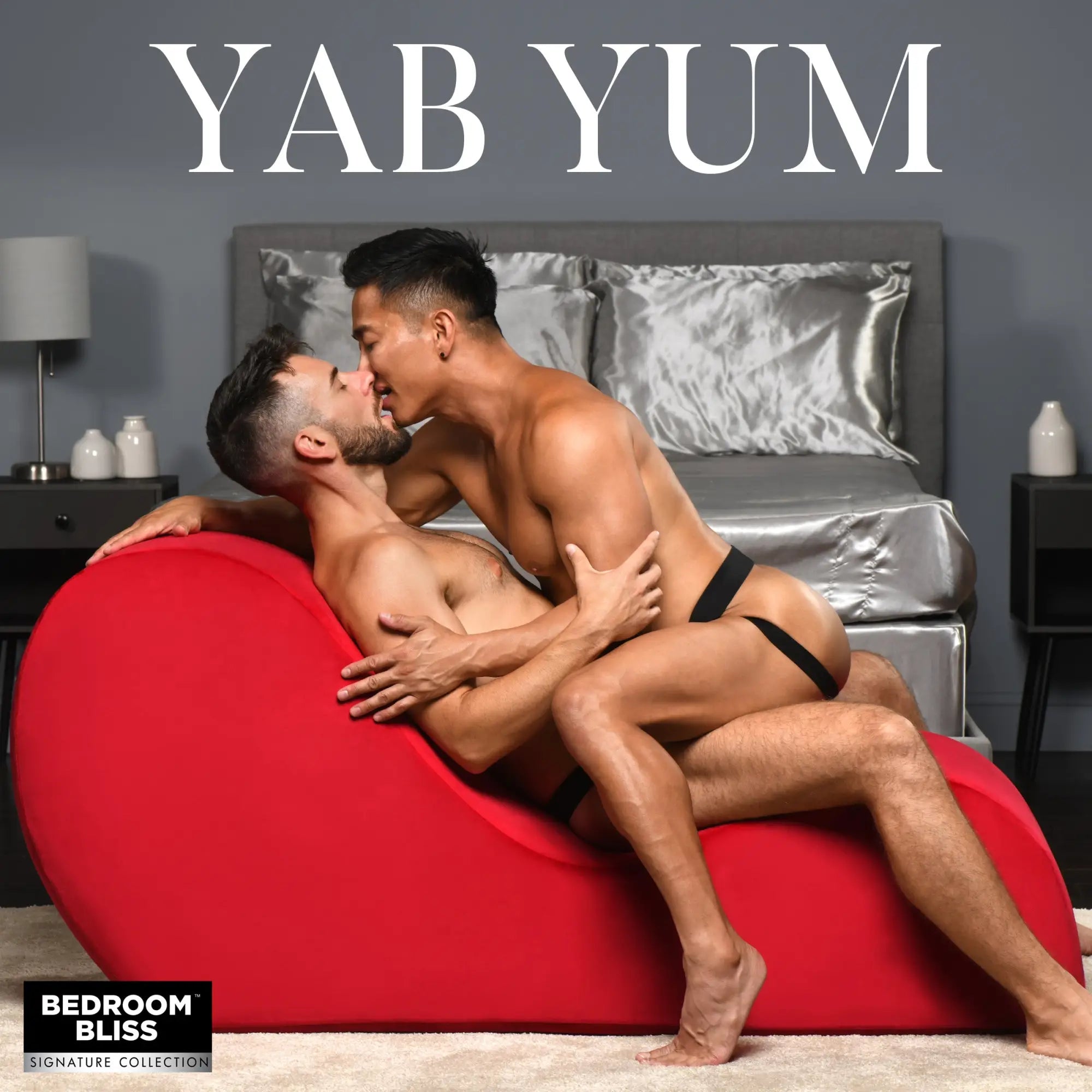 Two men kiss on a bright red, ergonomically curved Bedroom Bliss The Love Couch in a bedroom setting.