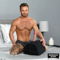Muscular man sitting on a bed with a black Bedroom Bliss Love Toy Cushion featuring an upright phallic attachment.