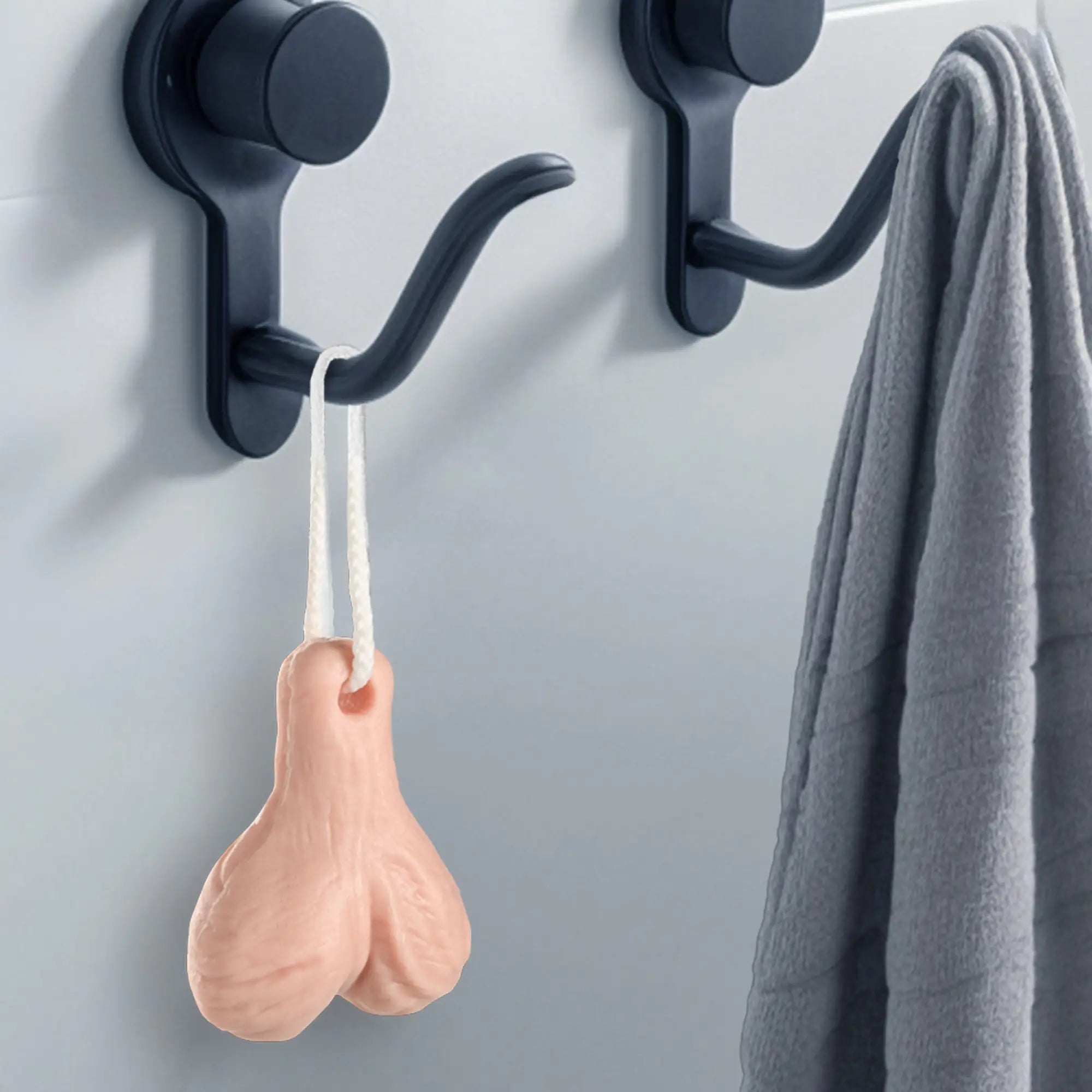 Big Ballers Balls Soap: A skin-toned, testicle-shaped soap hanging from a white string on a black bathroom wall hook.