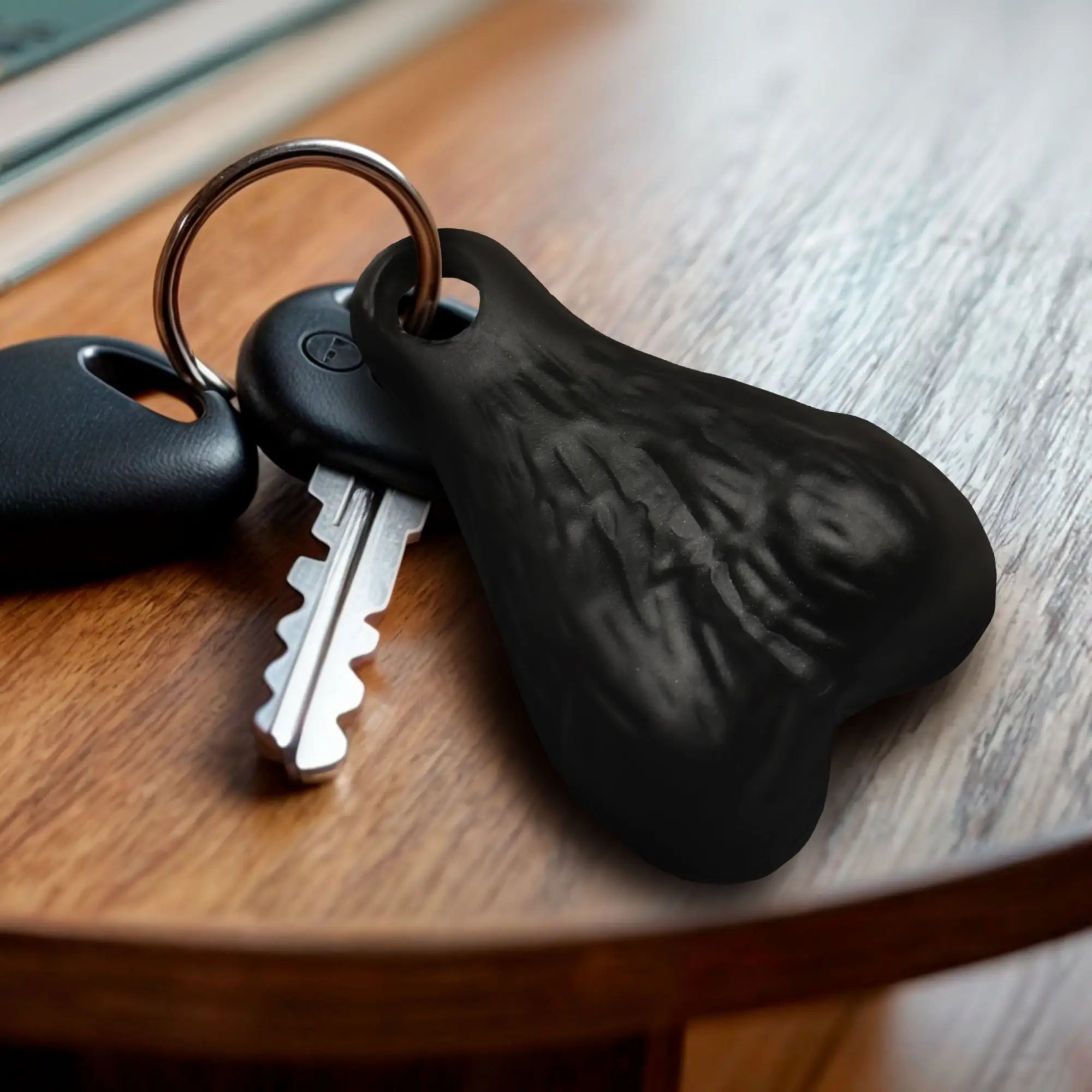 Big Ballers keychain: a textured, matte black, anatomical-shaped charm on a metal keyring with a car key on a table.