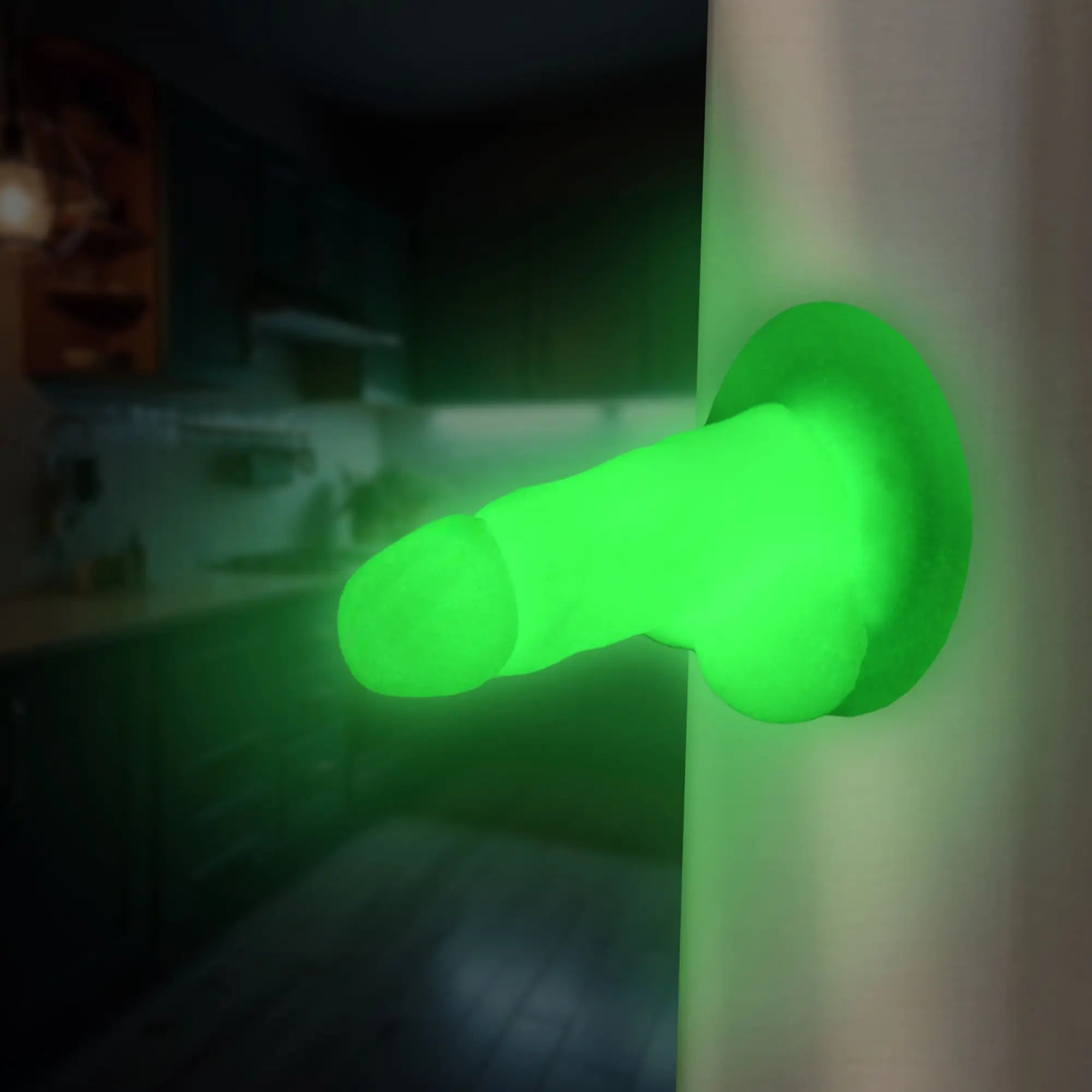 A bright neon green Big Ballers Suction Snap-On Pecker stuck to a surface in a dimly lit room.
