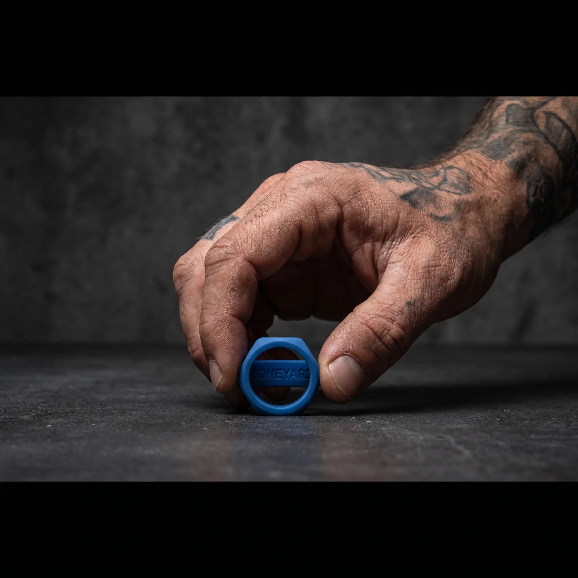 Boneyard Bust A Nut Blue hexagonal silicone cock ring on dark surface alongside a rugged man's hand