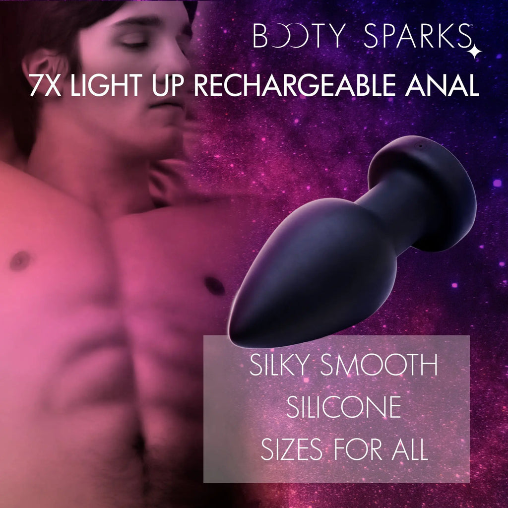 Booty Sparks 7X light-up, black silicone, plug-style anal toy against a purple and pink celestial background.