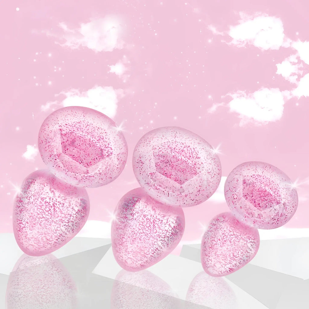 Booty Sparks Glitter Gem anal plug set: five sparkling, pink-tinted clear plugs set against a pink, cloudy background.
