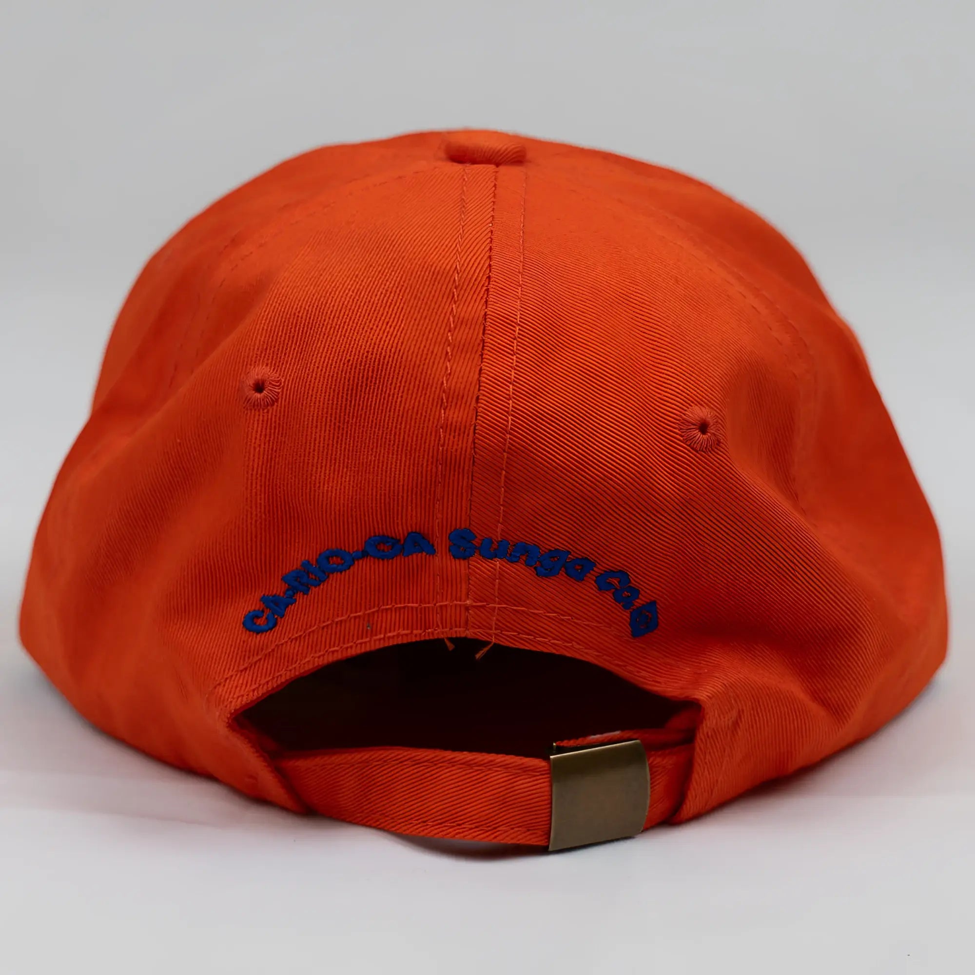 Back of bright orange ca-rio-ca crest logo embroidered designer dad hat with blue text and metallic buckle