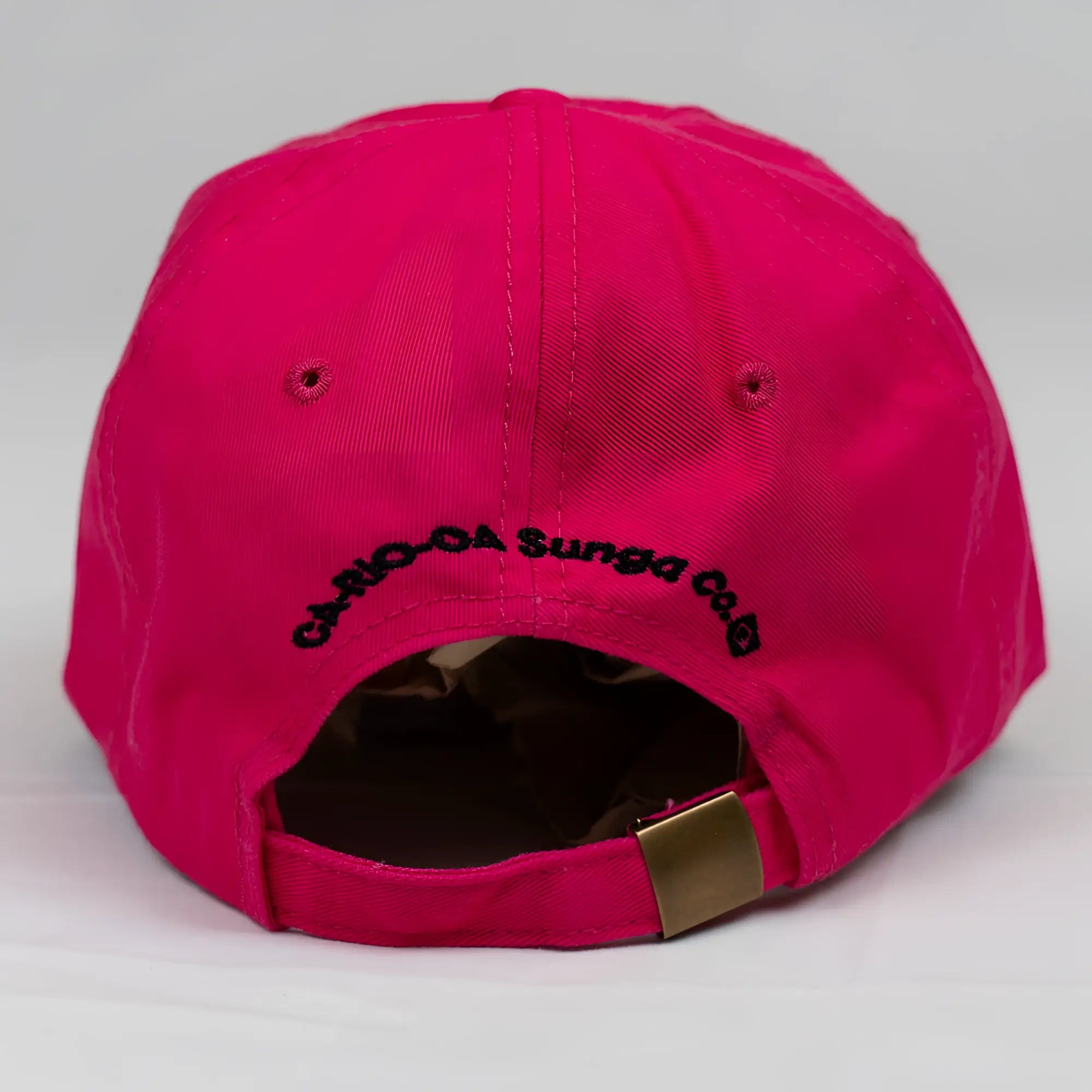The back side of a vibrant fuchsia ca-rio-ca crest logo embroidered designer dad hat with black text and gold-tone strap