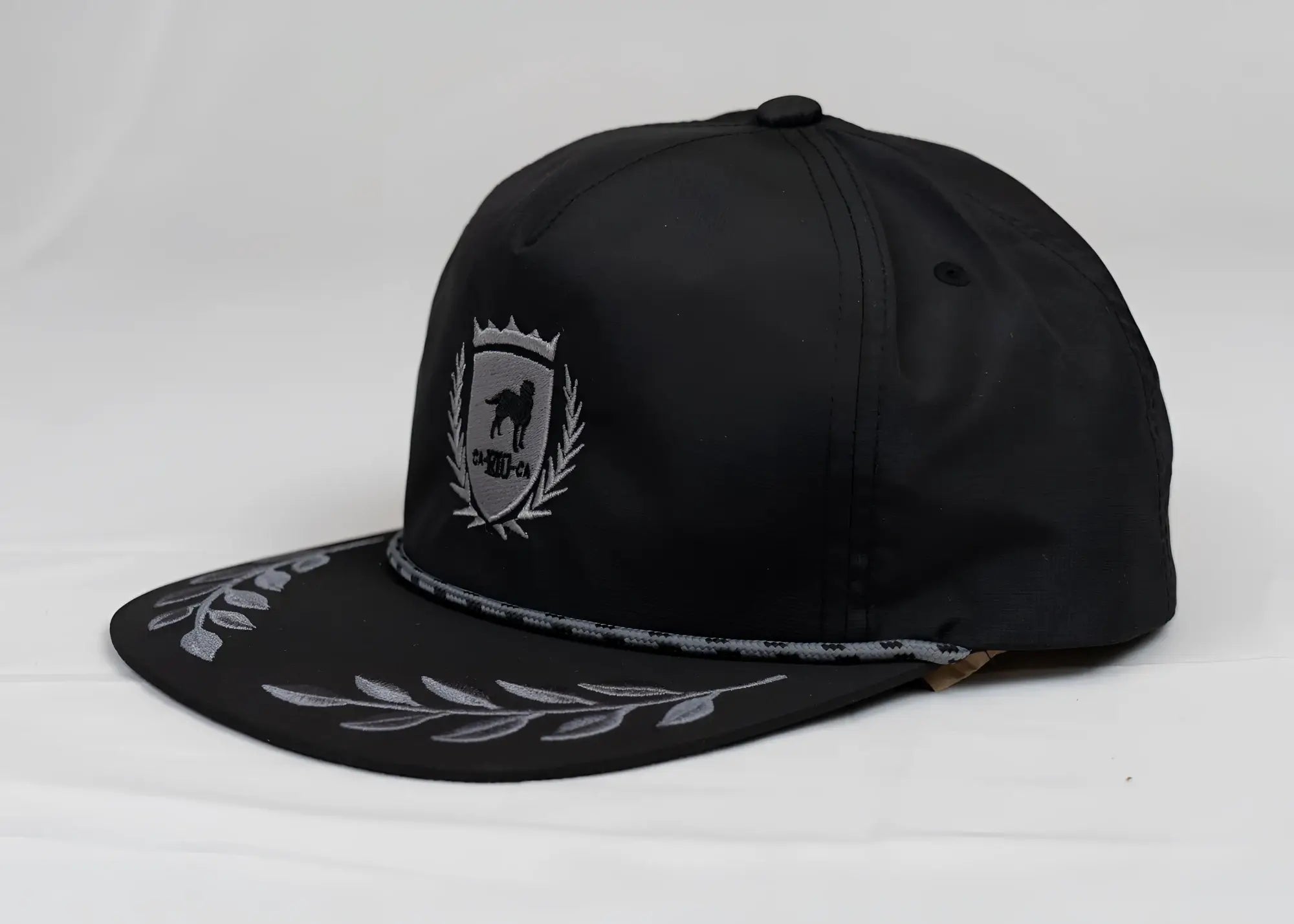 Black snapback cap with silver embroidered CA-RIO-CA crest logo and bay leaf pattern on the brim