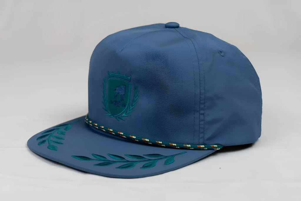 Blue snapback cap with teal embroidered CA-RIO-CA crest and leaf pattern on brim accented by braided trim