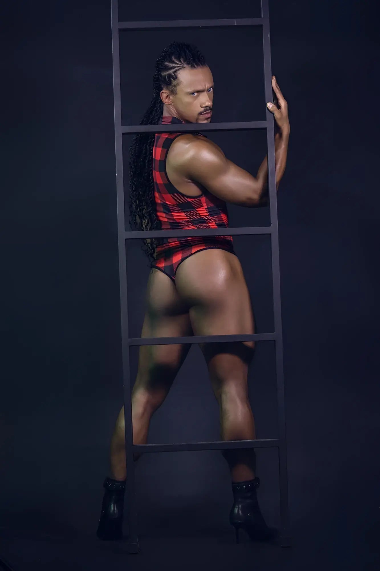 A muscular man with long braided hair wears a red and black plaid sleeveless CandyMan 99800 lumberjack bodysuit