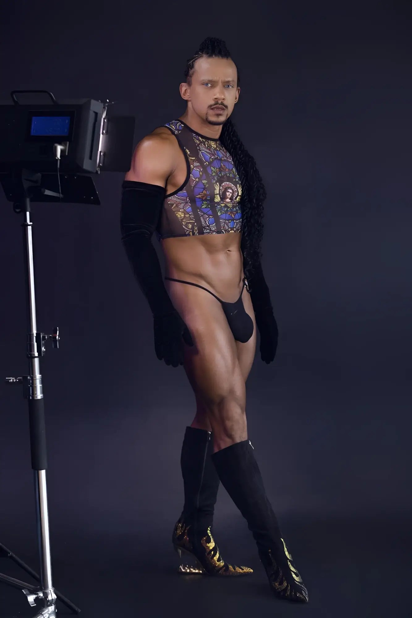 A man wearing a black patterned CandyMan 99805 stained glass tank top with sheer mesh and matching gold thigh-high boots